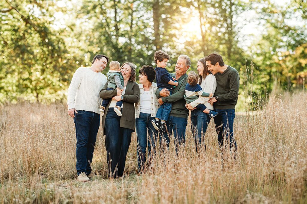 A group of three adults, three children, and an older couple stand together smiling and laughing in a sunlit, grassy field with trees in the background—perfect for Fall Family Photos in Massachusetts.