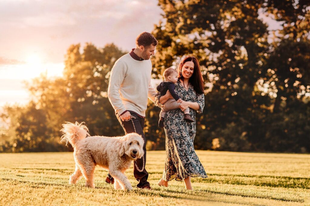 A man, woman, and baby walk barefoot on grass at sunset in Massachusetts, smiling and enjoying time together. The woman carries the baby, and a fluffy dog walks happily beside them—perfect for Fall Family Photos with trees and a golden sky behind.