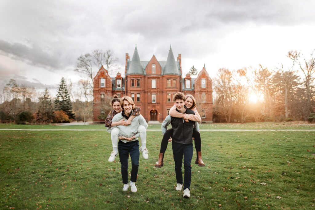 Two men are giving piggyback rides to two women on a grassy field in front of a large, castle like building in Massachusetts. The setting sun casts a warm glow, perfect for fall family photos. The group is smiling and dressed casually.