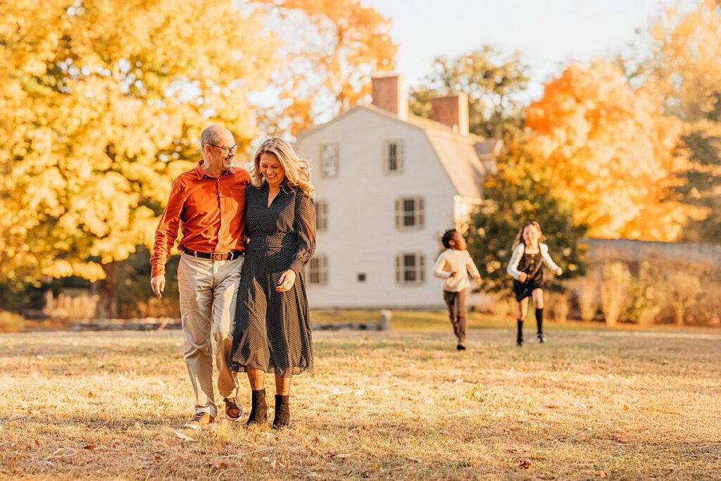 A smiling couple walks arm in arm across a sunlit field with autumn trees and a white house in the background, while two children run and play behind them—perfect for capturing Fall Family Photos in Massachusetts.