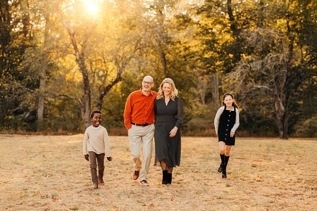 A family of four, two adults and two children, walk together in a sunny Massachusetts park. They are smiling and enjoying each others company, surrounded by golden leaves—perfect for Fall Family Photos.