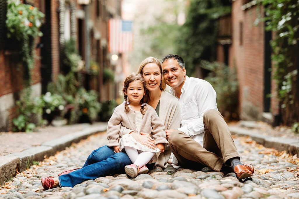A family of three sits happily on a cobblestone street in Massachusetts, surrounded by fall foliage. The parents are close together, smiling warmly, with their young child in front. A sense of warmth and invitation fills the scene, highlighted by an American flag waving gently nearby.