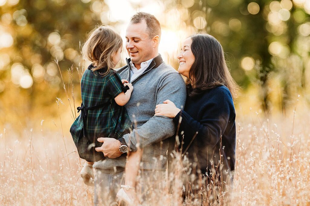 A man and woman smile while standing in a sunlit field, with the man holding a young girl in a green plaid dress. Tall dry grass and golden light surround them, capturing the warmth of Fall Family Photos in Massachusetts.