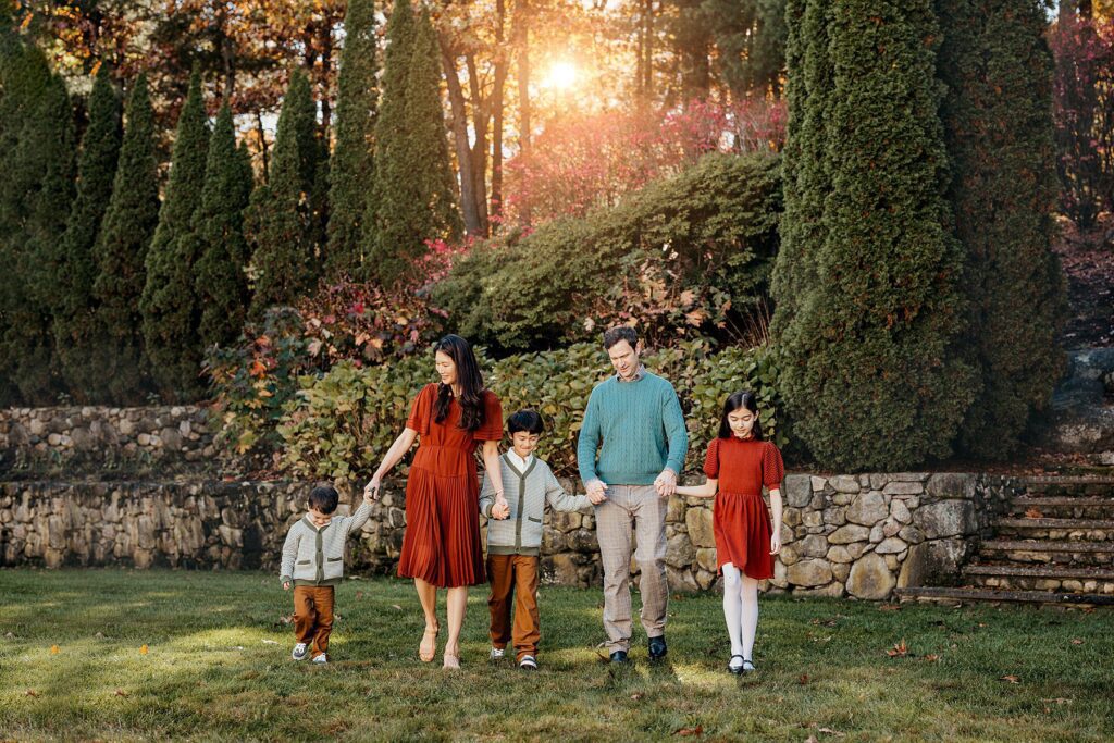 A family of five strolls hand in hand on the grass before a stone wall and tall trees with vibrant autumn foliage in Massachusetts. The parents stand at the center, each holding two childrens hands, as the warm glow of the setting sun enhances their Fall family photo backdrop.