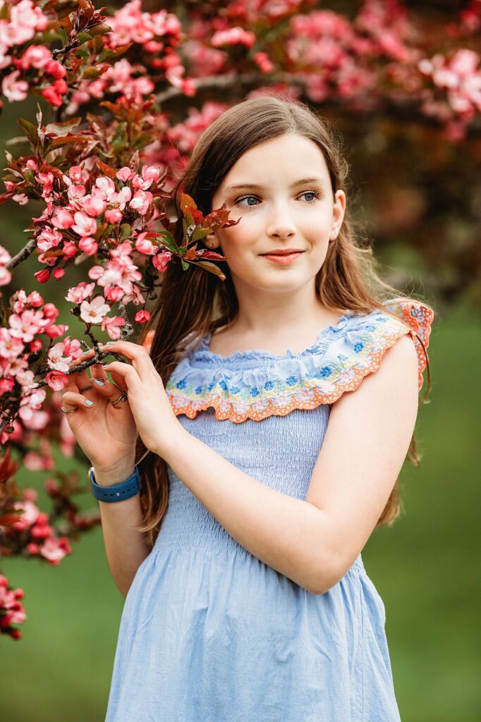 A girl in a blue dress stands beside a blossoming cherry tree, gently holding a branch. She gazes to the side, surrounded by Bostons vibrant greenery.