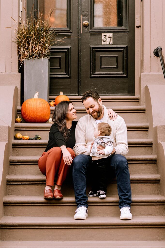 A family of three sits on brownstone steps decorated with pumpkins in Massachusetts. The parents smile at each other while holding their baby, who looks at the father. The scene is cozy and perfect for Fall Family Photos.