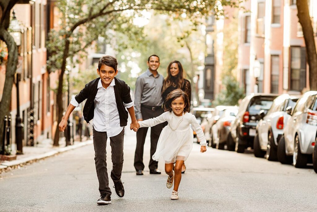 Two children, a boy and a girl, hold hands and run towards the camera on a tree lined Massachusetts street as two adults smile behind them. Parked cars line both sides—a perfect setting for Fall Family Photos.