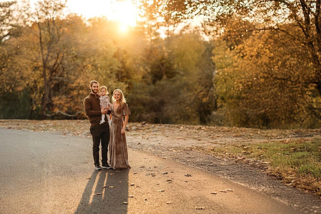 A smiling couple stands on a road with their young child at sunset, surrounded by autumn trees with golden leaves in Massachusetts. The sunlight casts long shadows behind them, making it a perfect scene for Fall Family Photos.