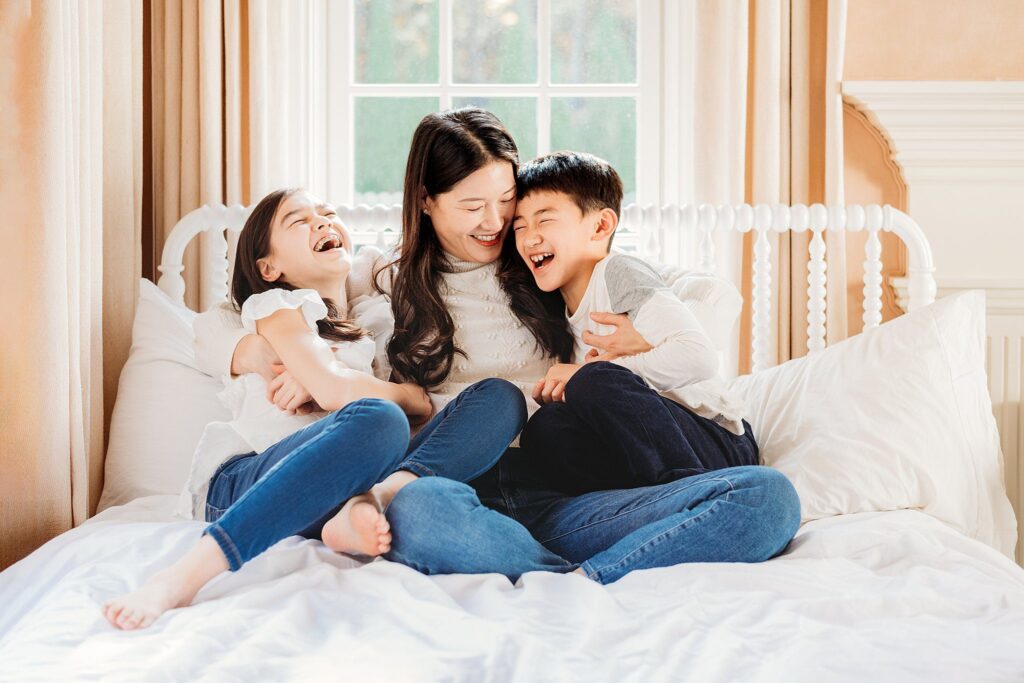 A woman and two children sit on a bed, laughing together. The woman is in the middle, embracing the children. Dressed casually, they bask in the warm glow of a bright window. The cozy scene captures a perfect moment for fall family photos in Massachusetts.