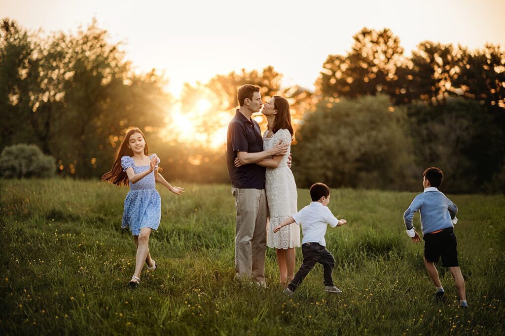 In a serene and joyful scene of spring family photography, a couple shares a kiss in a sunlit field at sunset while three children run and play around them, framed by the warm glow and trees in the background.