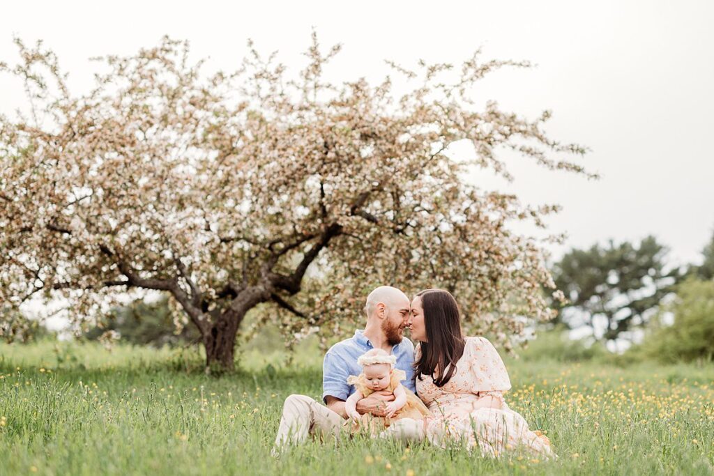 A family enjoys a spring session on green grass beneath a blossoming tree. A man and woman, sitting closely, lovingly touch foreheads as they hold their small child in a yellow outfit with a white headband. The overcast sky adds to the scenes peaceful ambiance.