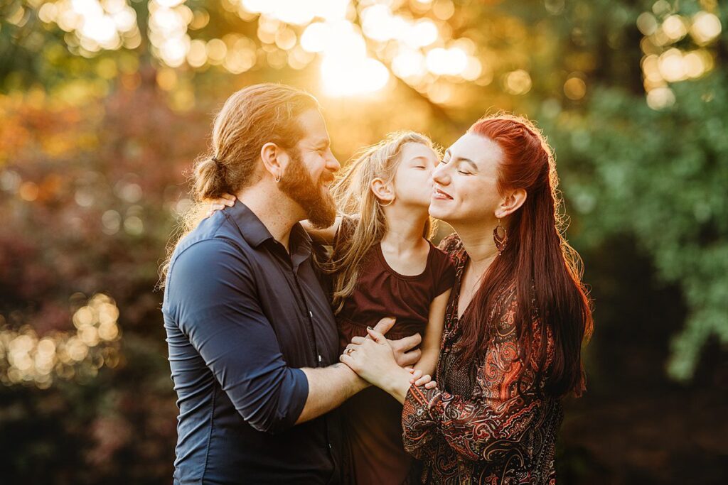 A family of three shares a loving moment during a Massachusetts sunset. The child is held by the parents, leaning in for a kiss with the mother. The sun casts a warm glow, highlighting the sunset in the background.