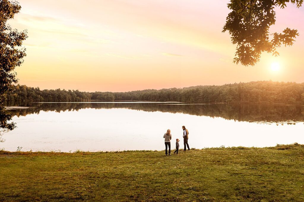 A family of four stands by a tranquil lake in Massachusetts at sunset, the sky painted in soft orange hues. Trees frame the sides, and the sun is low on the horizon, reflecting on the water. This moment epitomizes perfect fall family photography—peaceful and warm.