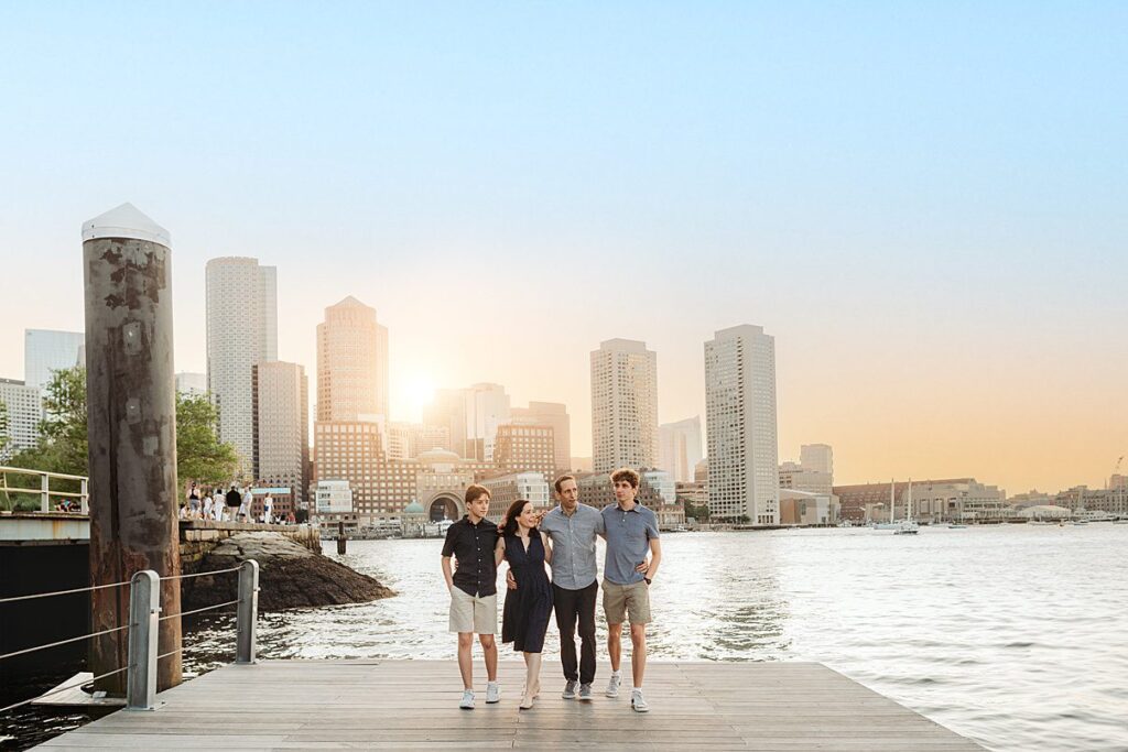 A family of four walks arm in arm on a dock by the water, with a city skyline and sunset in the background. Smiling together, they enjoy warm, relaxed moments—perfect for unforgettable Beach Sessions.