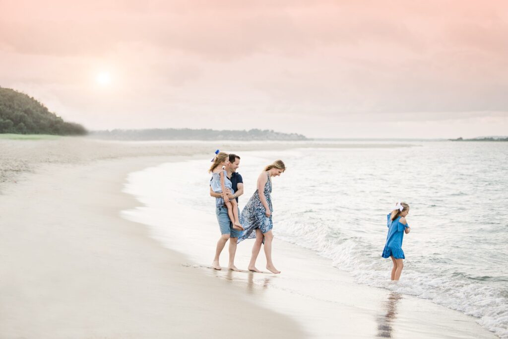A family of five enjoys Beach Sessions at sunset, strolling along the sandy shore. Two adults and two children walk together while another child in a blue dress stands at the calm water’s edge beneath a pink sky.