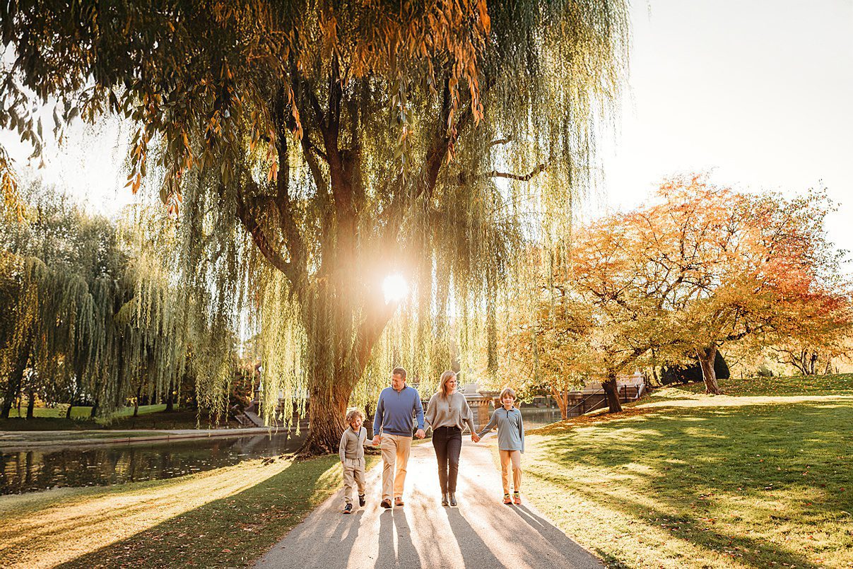 A family of five walks hand in hand on a sunlit path in a park, surrounded by green grass, autumn colored trees, and a large willow tree—one of the best fall family photo locations. Sunlight shines through the trees, casting long shadows.