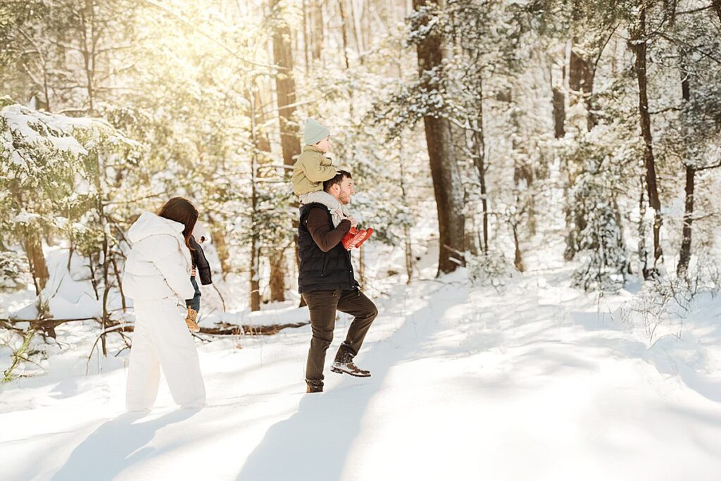 A family walks through a snowy forest for magical portraits. An adult carries a child on their shoulders while another walks nearby, all bundled in winter clothing, capturing the warmth of Boston winter family photos under sunlit trees.