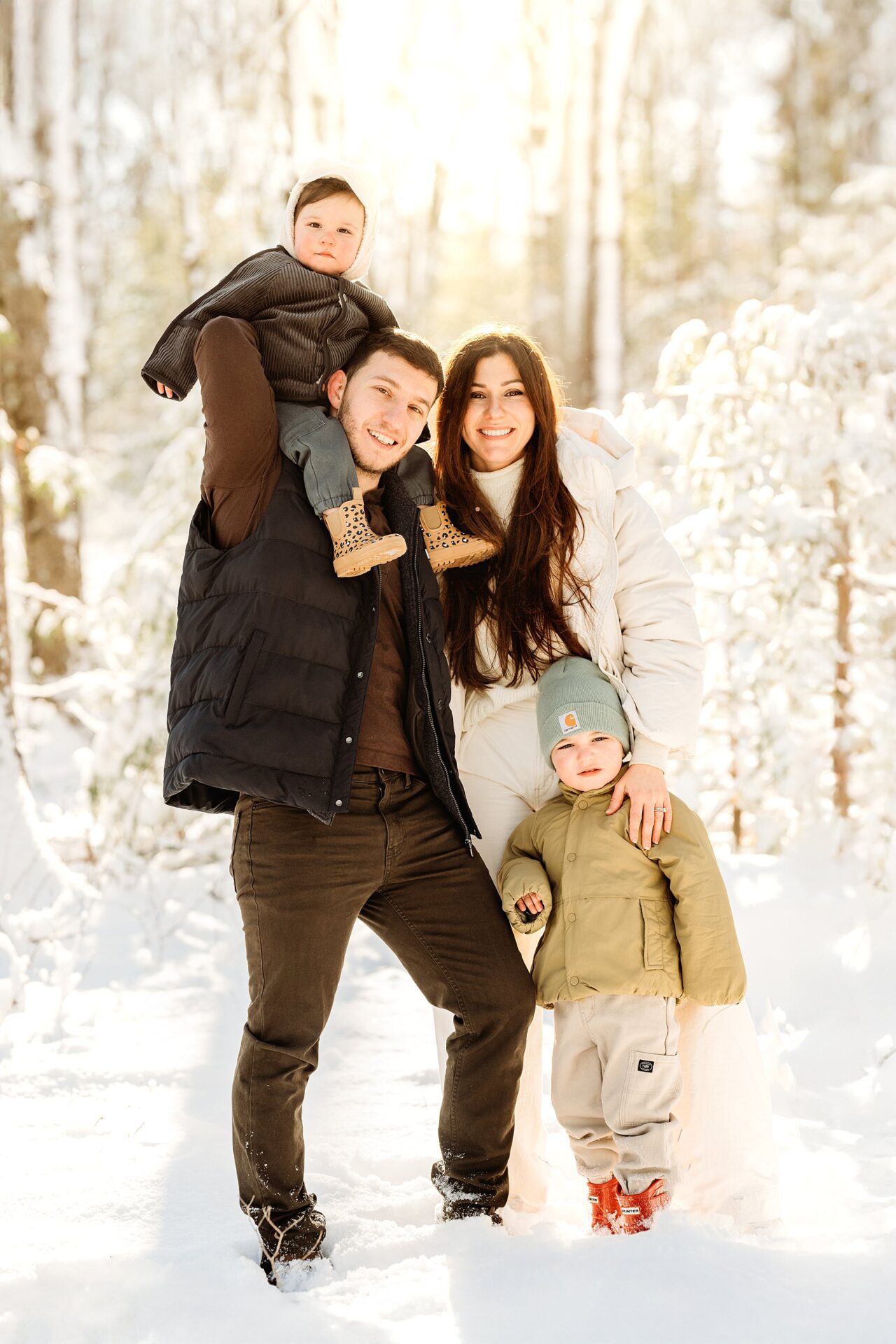 A family of four poses in a snowy forest for magical portraits. The father holds a baby on his shoulders while the mother stands beside him, holding their child's hand. They are all dressed warmly and smiling, capturing unforgettable Boston winter family photos.