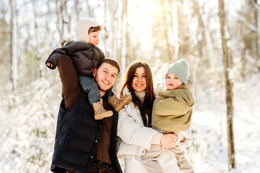 A smiling couple stands outdoors in a snowy forest, each holding a young child dressed in winter clothing. Sunlight filters through the trees, capturing the joy of magical winter photography and creating bright, cheerful snowy family portraits.