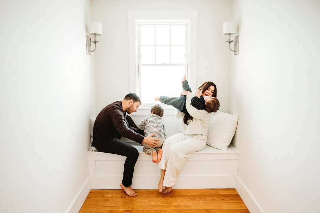 A family of four sits in a bright nook by a window. The father playfully holds one child, while the mother laughs and hugs the other. Everyone appears joyful and relaxed during their cozy Boston winter family photos.
