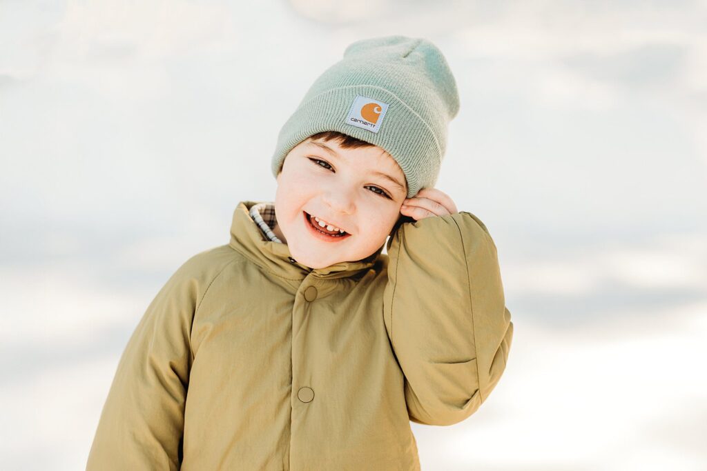A young child wearing a light green beanie and a tan winter coat smiles cheerfully outdoors, capturing the joy of snowy sessions against a bright, wintry backdrop.