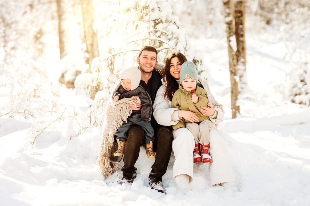 A family of four sits together on a snowy log in a sunlit winter forest, bundled in warm clothes. The parents smile at the camera while holding their young children, creating magical portraits perfect for Boston winter family photos.