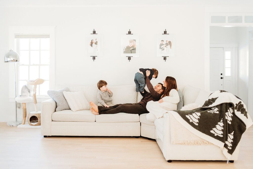 A family of four relaxes on a white sectional sofa in a bright living room; above them, magical portraits and Boston winter family photos hang on the wall as one parent lifts a toddler and the other sits with a young child.