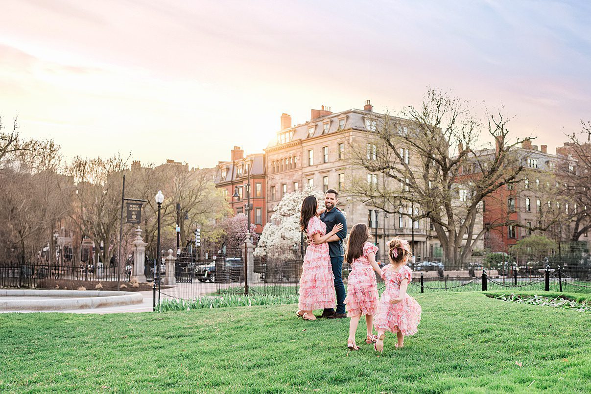 A family of five stands on a grassy lawn at sunset, with historic buildings in the background—one of the best spring family photo locations. The mother and three daughters wear matching pink dresses, while the father stands behind them, holding the youngest child.