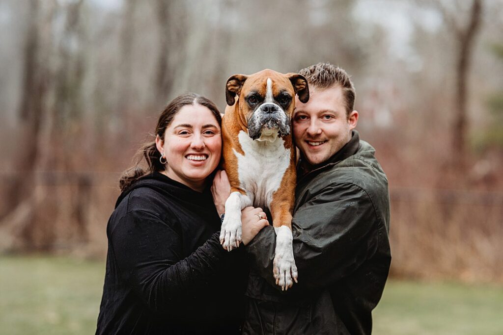 A woman and a man are standing outdoors, smiling and holding a brown and white boxer dog between them. The background is blurred with trees and grass, capturing a natural, relaxed Family Photography moment.