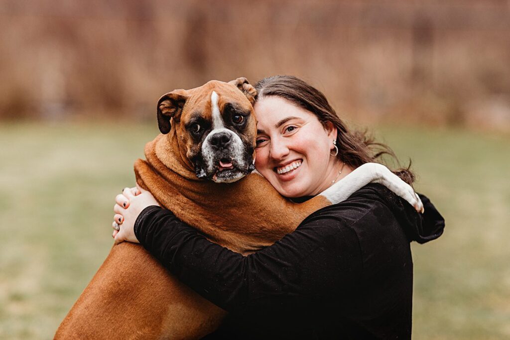 A woman smiles while hugging a large brown and white boxer dog outdoors. Captured by a Boston Dog Photographer, the pair look happy and close as they embrace, framed by blurred greenery in this heartwarming family photography moment.