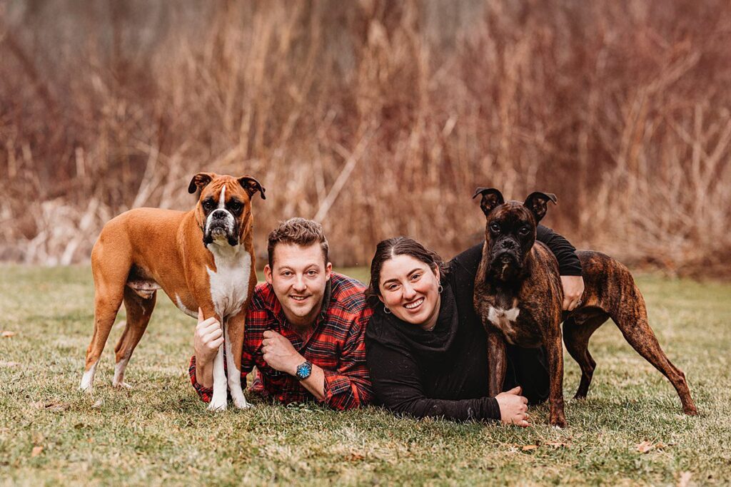 A man and woman lie on the grass with their dogs, capturing a candid moment of family photography.