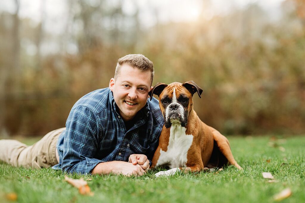 A man in a blue plaid shirt lies on the grass next to a brown and white boxer dog. Both appear relaxed, looking at the camera—a perfect moment of family photography captured by Helena Goessens Photography.