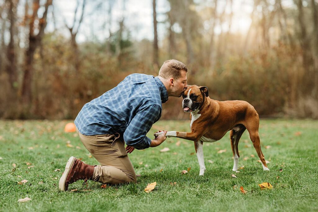 A man kneels on grass, holding his brown and white dog's paw. He kisses the dog's head, outdoors in a park with scattered leaves—capturing a heartfelt moment by Helena Goessens Photography