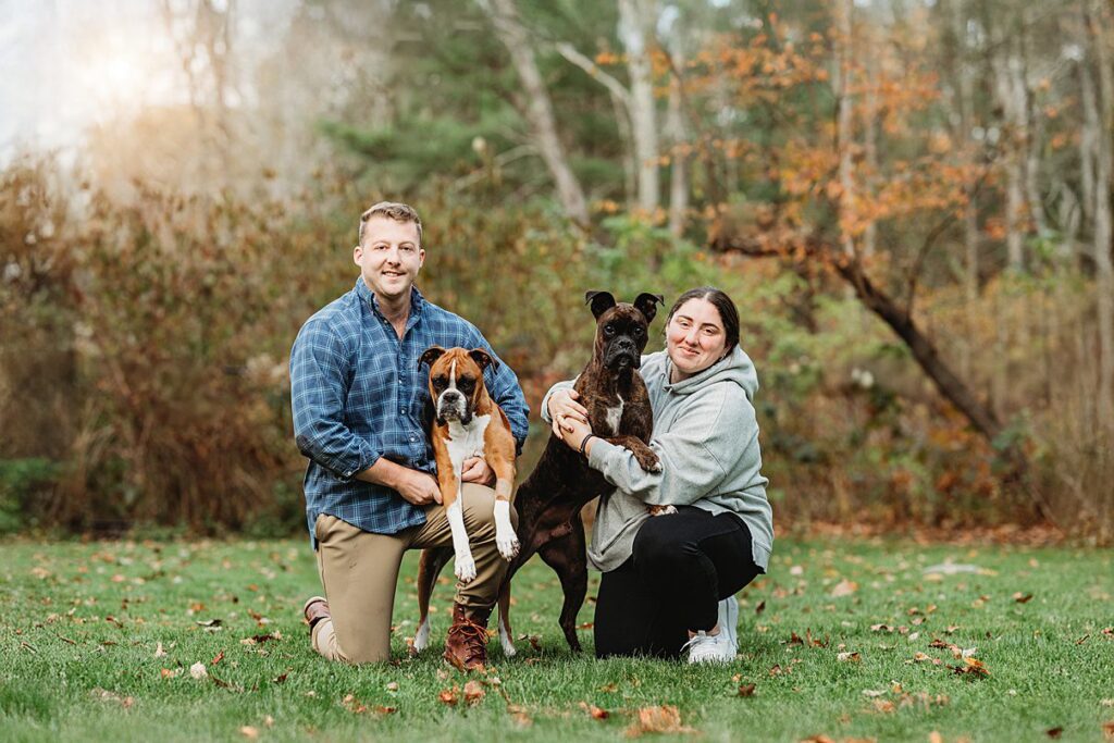 A man and a woman kneel on grass outdoors, each holding a dog, smiling at the camera. Autumn trees with orange leaves set the scene—perfect for a Dog and Family Photographer capturing joyful moments of family photography.