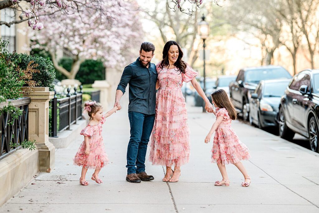 A family of four stands on a city sidewalk lined with blooming trees, smiling and holding hands. The mother and two young daughters wear matching pink dresses, while the father wears a blue shirt—capturing perfect Spring Family Photos in Boston.