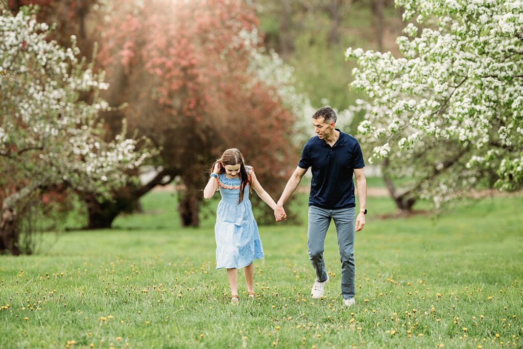 A man and a young girl holding hands walk through a green grassy field with blooming trees, both smiling. The scene is bright and spring like, with the girl in a blue dress and the man in navy and gray.