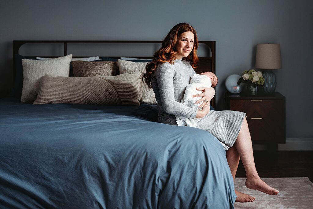 A woman with long red hair sits on the edge of a neatly made bed, holding a swaddled newborn in her arms. She smiles softly in a cozy bedroom with blue and neutral tones—an intimate moment captured through in home newborn photography.