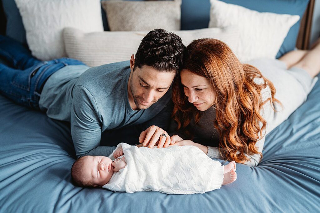 A man and woman lie on a bed, looking lovingly at their swaddled newborn baby who is sleeping. The parents’ hands gently rest near the baby, capturing a tender in home newborn photography moment with soft pillows in the background.