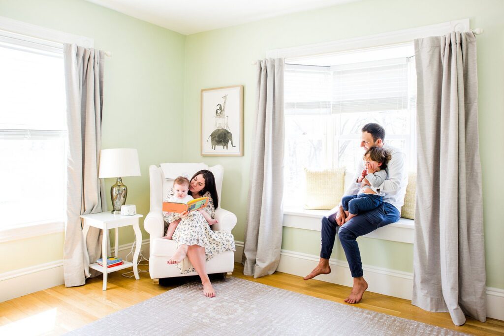 A woman sits in a white armchair reading to a small child, while a man on a window seat holds another. The bright room with light green walls, large windows, and gray curtains.