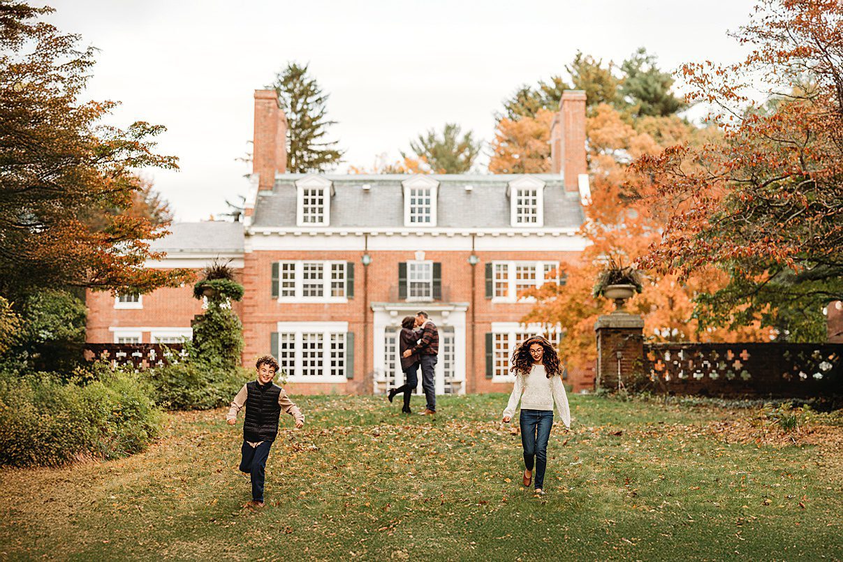 Two children run across a grassy lawn toward the camera, while a couple embraces in the background in front of a large brick mansion surrounded by autumn trees.