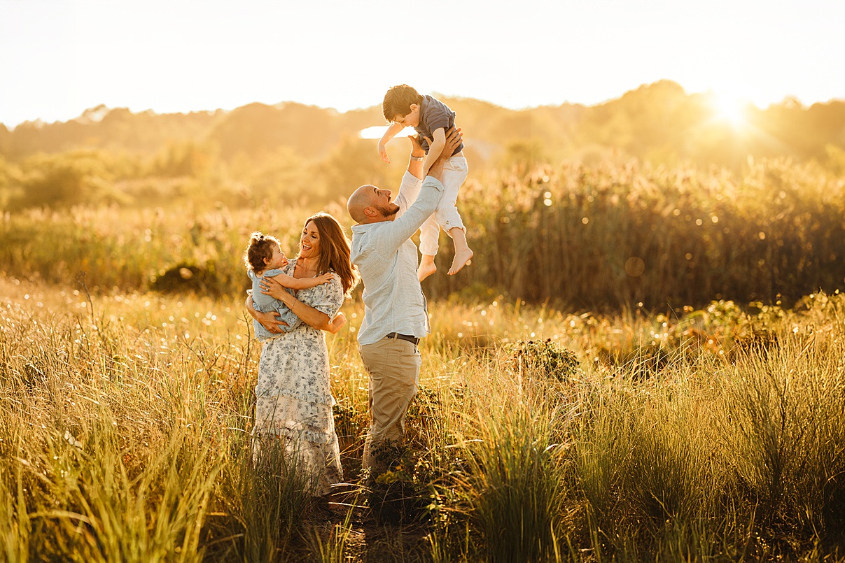 A family of four stands in a sunlit field, much like the joy found in Beach Sessions. The mother holds a young child while the father lifts another into the air, surrounded by tall grass touched by warm sunlight.