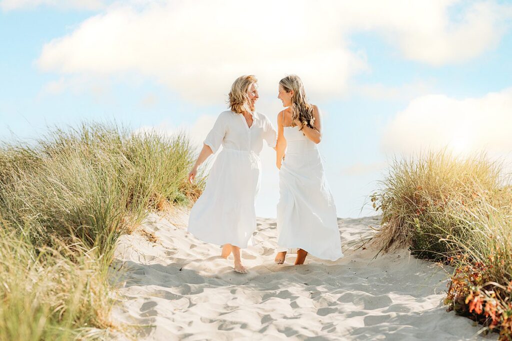 Two women in white dresses walk barefoot on a sandy path through tall grass, smiling at each other under a bright blue sky with fluffy clouds—capturing moments perfect for Family Photography Packages Explained.