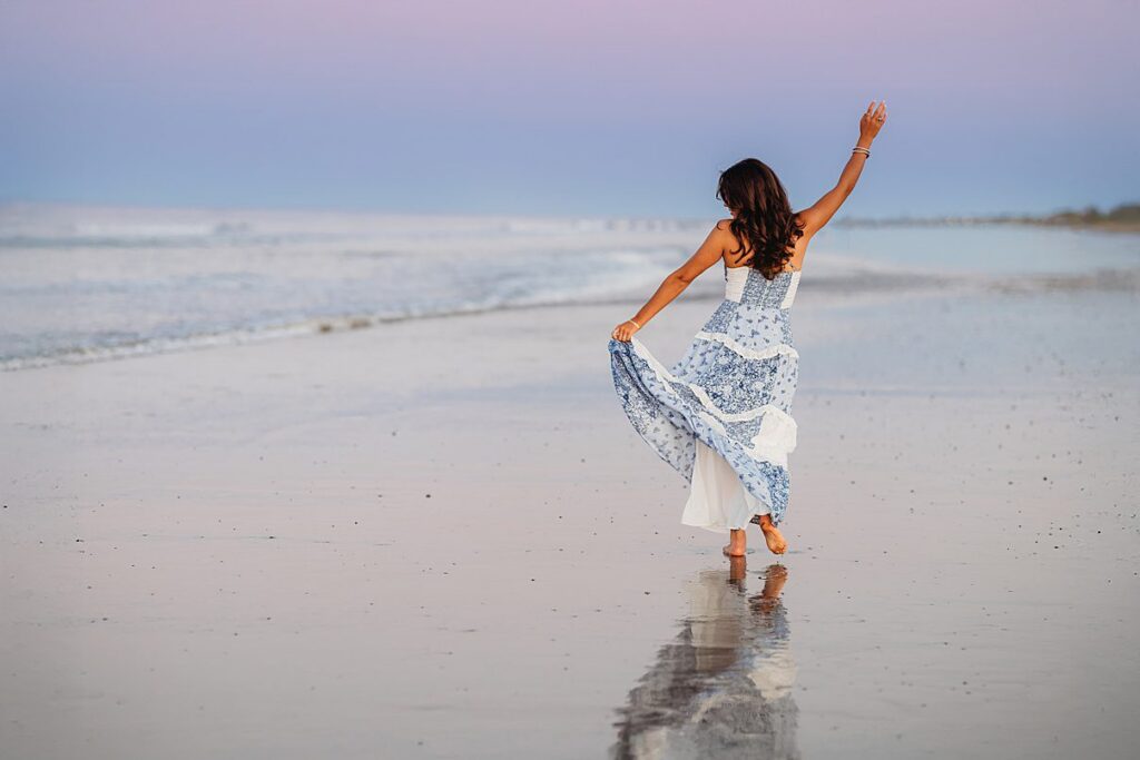 A woman in a flowing blue and white dress walks barefoot along a beach at sunset, holding up her skirt with one hand and raising the other—her reflection visible on the wet sand, showcasing moments captured with Family Photography Packages Explained.