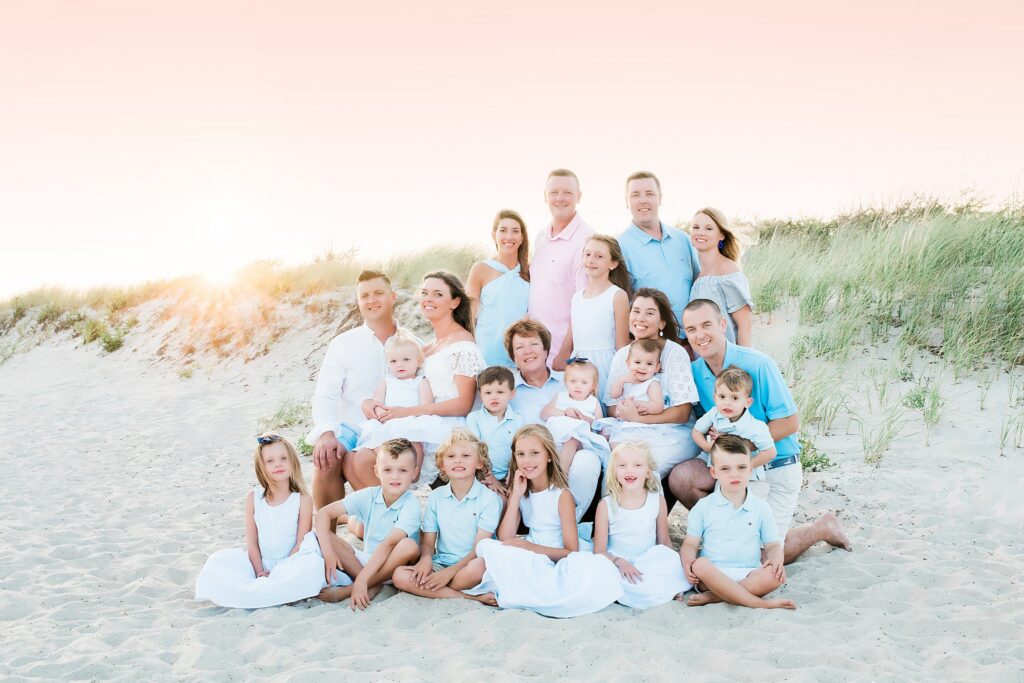 A large family poses together on a sandy beach at sunset, smiling in coordinated white and light blue outfits, with grassy dunes and a soft pink sky in the background—showcasing moments captured with Family Photography Packages Explained.
