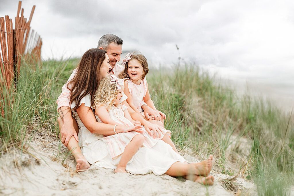 A family of four sits together on sandy dunes during their Beach Sessions, surrounded by tall grass. The parents and two young daughters are smiling and wearing light, summery clothes under a cloudy sky.