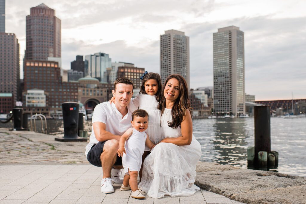 A family of four poses by the waterfront with tall city buildings in the background. Dressed in white, they smile and kneel together on a stone walkway near the water, capturing joyful memories during their Beach Sessions.