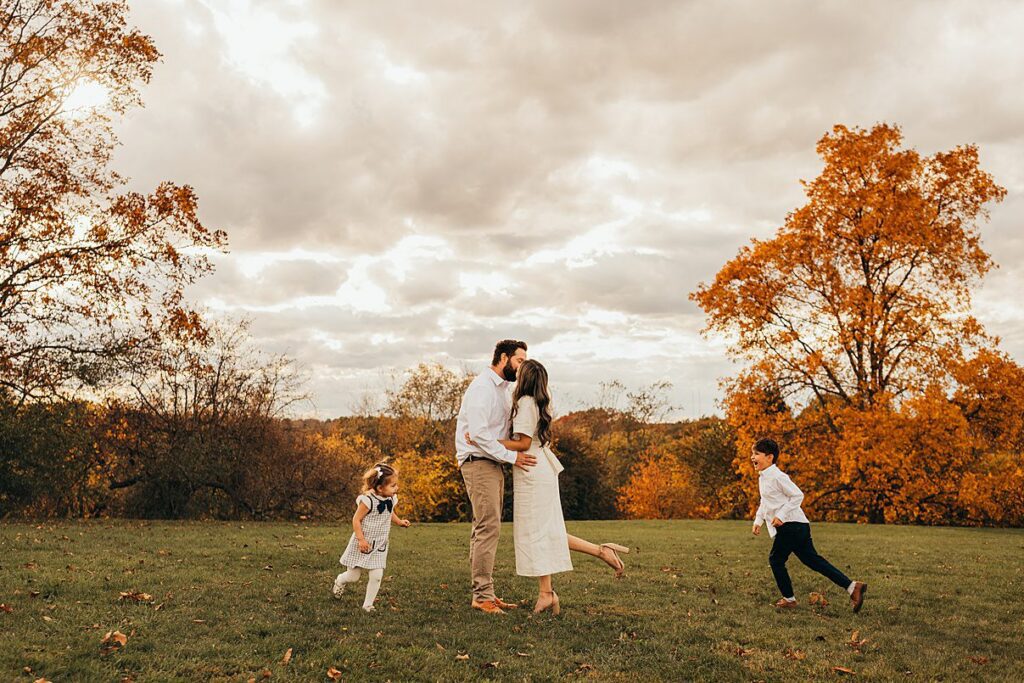 A couple kisses in the center of a grassy field with autumn trees, while two young children run and play around them under a cloudy sky—Family Photography Packages Explained capture moments like these beautifully.