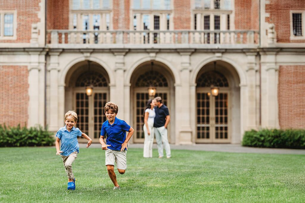 Two young boys run across a grassy lawn, smiling, with a large brick building in the background. A man and woman, slightly blurred, stand together—capturing the warmth of family moments perfect for Family Photography Packages Explained.