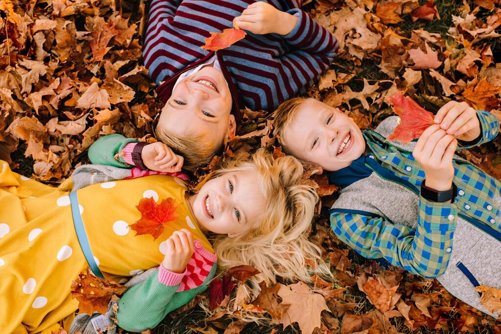 Three smiling children lie on colorful autumn leaves, each holding a bright red leaf, illustrating how to prepare kids for a photoshoot through playful, natural interaction. A girl in a yellow dress is nestled between two boys in colorful shirts, all looking up happily and capturing the joy of a successful family photoshoot.