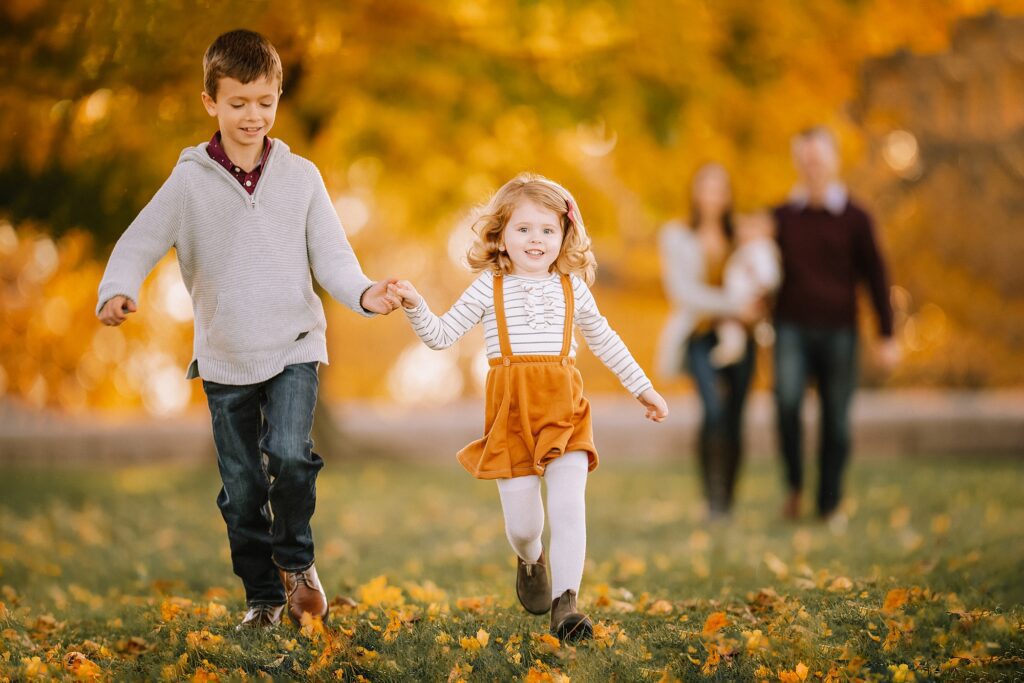 A young boy and girl run hand in hand across autumn leaves, smiling with joy during a family session, illustrating how to prepare kids for a photoshoot. Two adults and a baby stand softly blurred in the background among vibrant fall foliage, capturing the warmth, connection, and genuine moments of family photography.